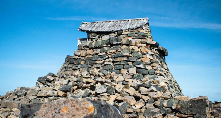 Steingebautes Gipfelschutzhütte auf einem Haufen eckiger Felsen unter einem klaren blauen Alpenhimmel.