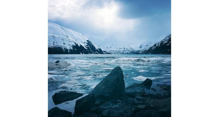 Icy lake with snow-covered mountains under a cloudy sky.
