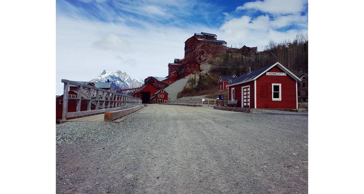Historic red buildings next to a gravel road with mountains in the background.
