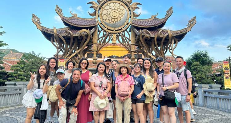 Grote groep mensen die poseren voor een decoratieve structuur in een zonnige omgeving.