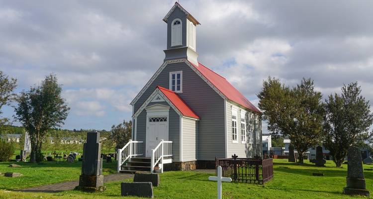 Petite église blanche avec un toit rouge dans une cour herbeuse sous un ciel nuageux.