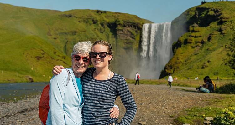 Two people posing in front of a waterfall and green hills.