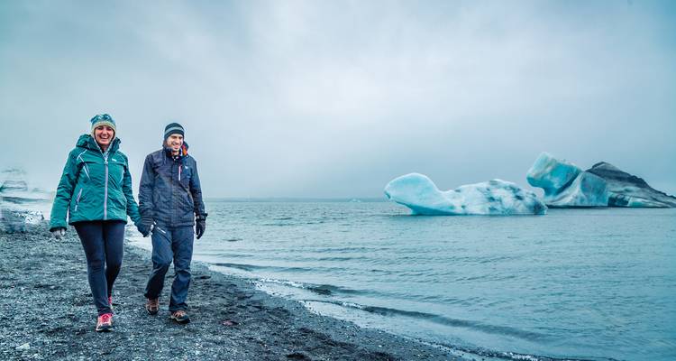 Two people walking by the sea with icebergs in the distance.