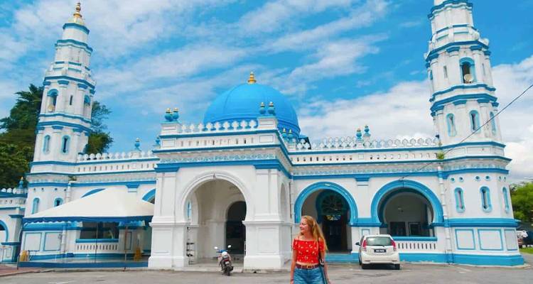 Mezquita azul y blanca con dos minaretes gemelos bajo un cielo brillante, turista de pie al frente.