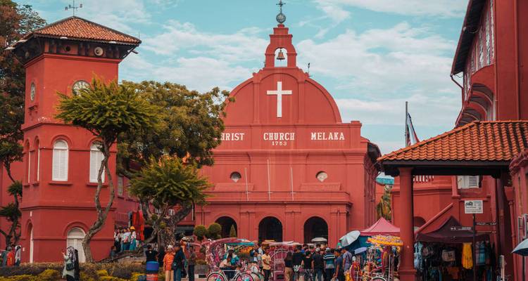 Icónica iglesia de Cristo pintada de rojo en Melaka con multitudes y rickshaws de bicicleta en la plaza.