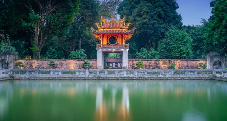 Estructura arquitectónica asiática iluminada junto a un estanque tranquilo y vegetación.