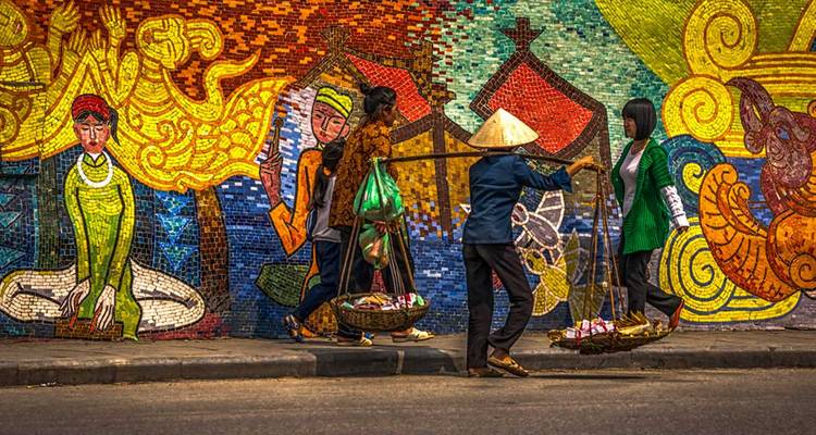 Escena callejera con paredes de mosaico vibrante y personas cargando mercancías.