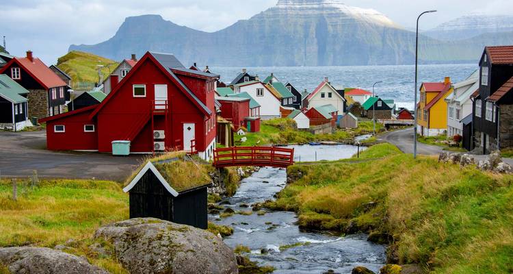 Casas coloridas de un pequeño pueblo de las Islas Feroe junto a un arroyo con acantilados marinos imponentes detrás.