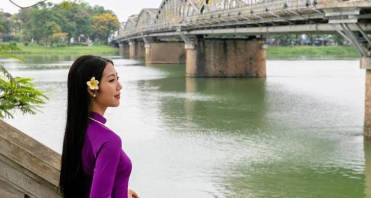 Young woman in purple traditional dress with a flower in her hair gazing across a calm river toward a bridge