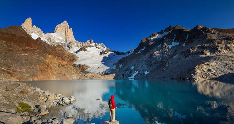 Personne en veste rouge debout près d'un lac bleu immaculé avec des montagnes.