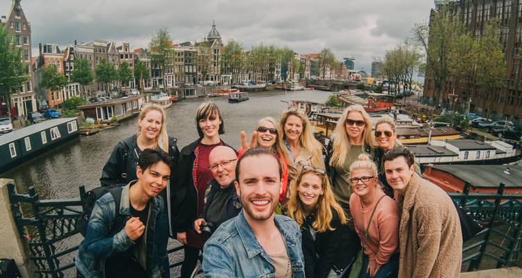 Selfie joyeux d'un groupe sur un pont enjambant les canaux d'Amsterdam, avec des maisons historiques et des péniches derrière eux par un jour nuageux.