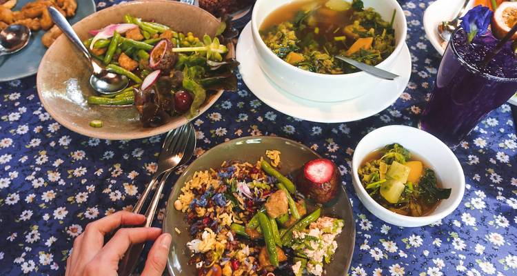 Variados platos tailandeses dispuestos sobre un mantel floral con una mano extendiéndose