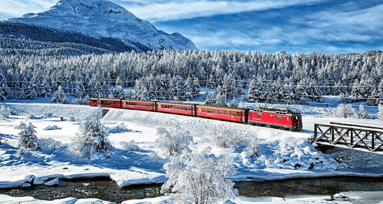 El tren rojo serpentea a través de un paisaje alpino nevado con bosque denso y congelado, y un pico imponente bajo la luz invernal.