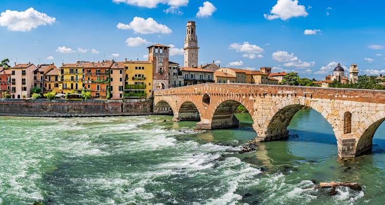 Un puente de arco de piedra histórico se extiende sobre un río verde y de rápida corriente con coloridos edificios de Verona y una torre más allá.