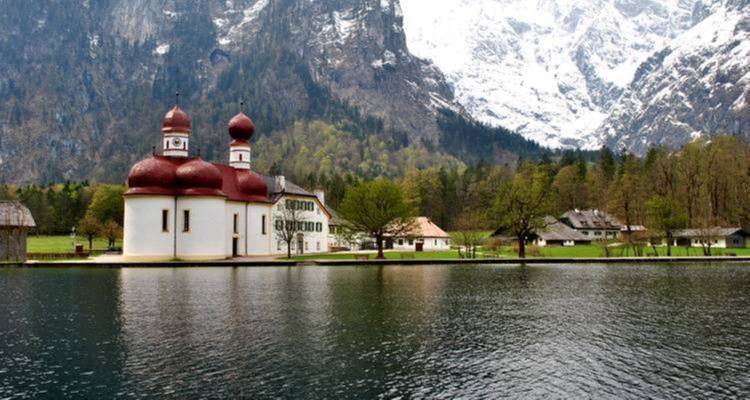Vista panorámica de una capilla junto al lago con montañas cubiertas de nieve.