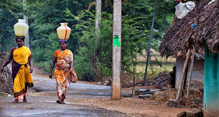 Two women in traditional attire carrying pots on their heads in a rural setting.
