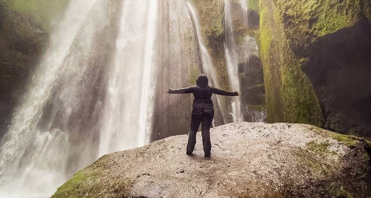 Un aventurier se tient les bras étendus sur un rocher moussu sous une puissante cascade dans une gorge islandaise brumeuse