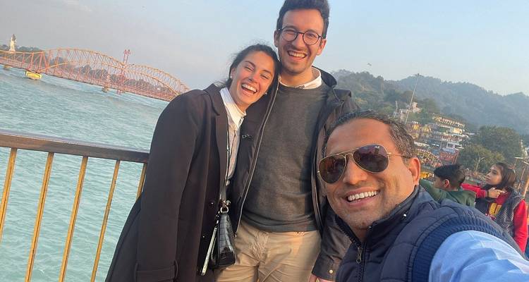 Three people smiling near a river with a bridge and buildings in the background.