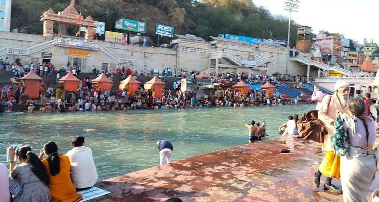 Many people at the ghats by a river with temples in the background.