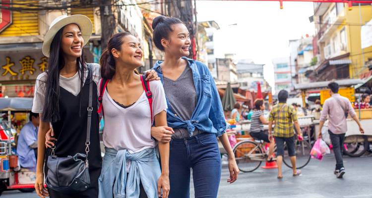 Drei Frauen gehen eine belebte Straße mit Geschäften und Schildern entlang.