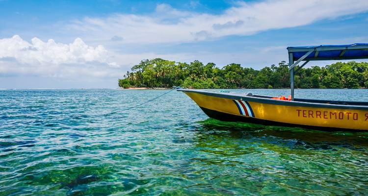 A small yellow motorboat anchored on crystal-clear turquoise water, with a lush green island in the distance under a bright sky.