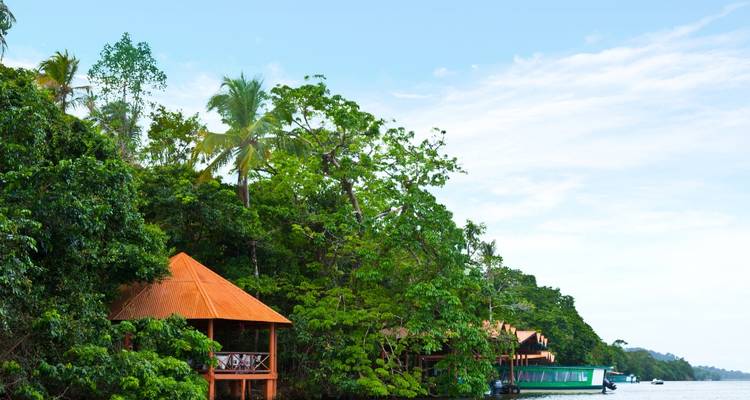 Jungle lodges with orange roofs line a lush riverbank below a blue sky.