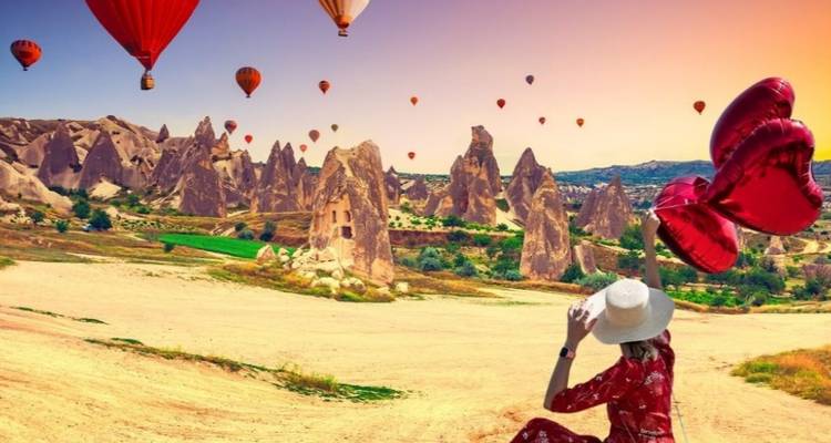 Woman holding red heart balloons in a hot air balloon field at sunset.