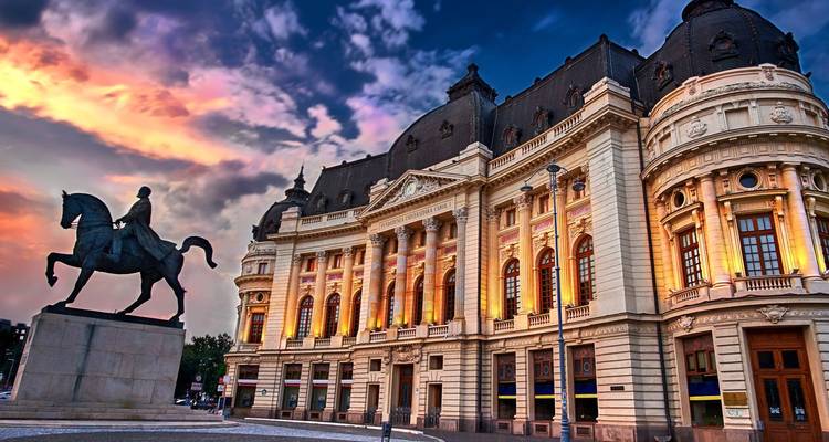 Elegant neoclassical Central University Library in Bucharest glowing at sunset with dramatic colorful clouds overhead and an equestrian statue in front.