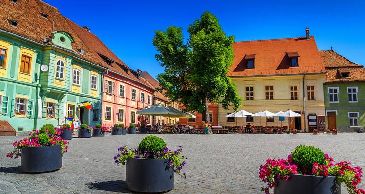 Brightly painted medieval houses surrounding a quaint square in Sighisoara with colourful flower pots under a vivid blue sky.