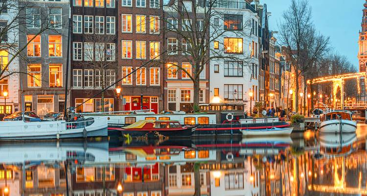 Amsterdam canal houses glow at dusk, reflecting perfectly in still water alongside moored boats.