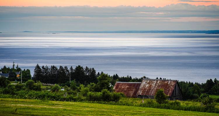 Graneros rústicos en una ladera cubierta de pasto que dan hacia un amplio y tranquilo estuario al atardecer.