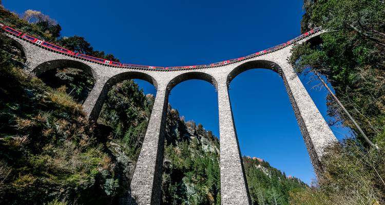 Train rouge circulant sur un haut viaduc en pierre dans une zone forestière.