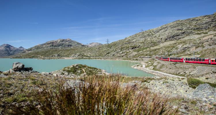 Train rouge voyageant le long d'un lac turquoise avec des montagnes.