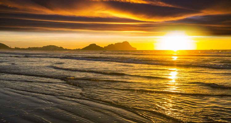 Vue du coucher de soleil sur une plage avec des vagues et la silhouette d'une île rocheuse.