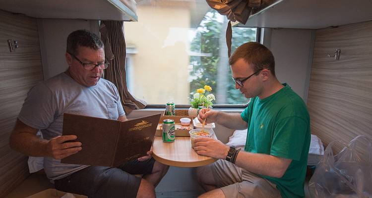 Deux hommes dînent dans une cabine de train. L'un lit un menu, et l'autre mange des nouilles instantanées.