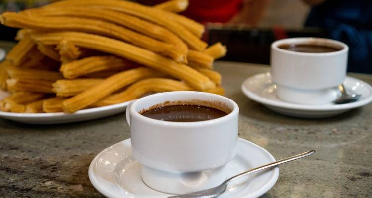 Two white cups of chocolate next to a stack of churros on a table.