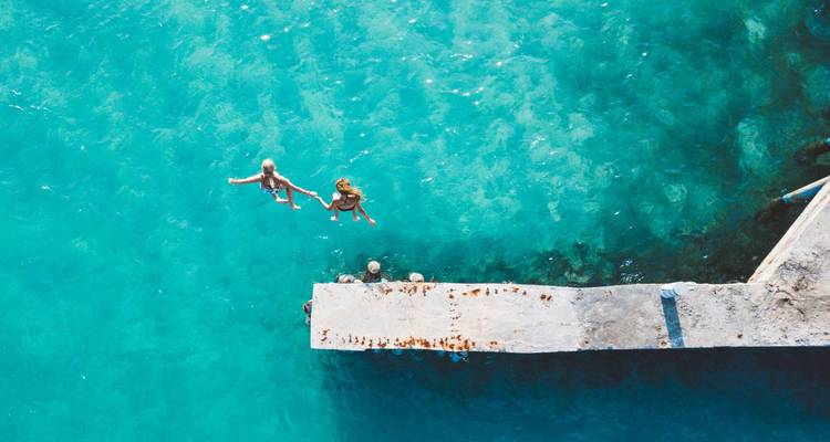 Aerial view of two people swimming in clear blue water near a concrete pier.