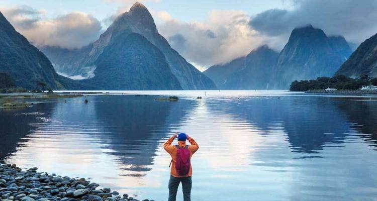 Un viajero fotografía los picos brumosos de Milford Sound reflejados en las tranquilas aguas del fiordo al amanecer.