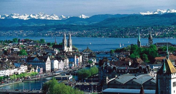 Paysage urbain avec une rivière, deux clochers d'église et des montagnes.