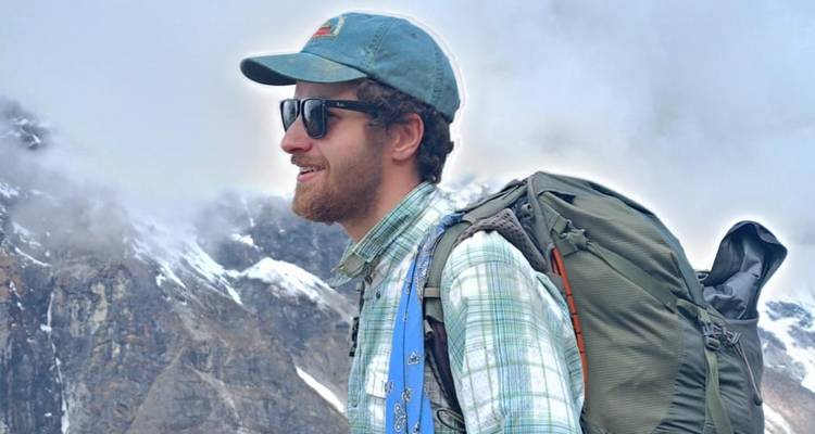 Man wearing a hat and backpack in front of snowy peaks.