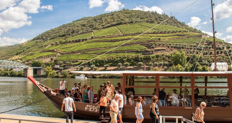Boat with people on a river, vineyard hills behind.