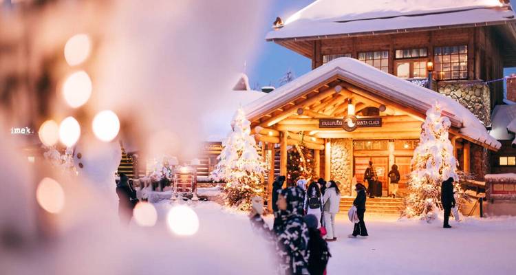 Cabaña de madera cubierta de nieve iluminada por luces cálidas con visitantes disfrutando del Pueblo de Papá Noel, Rovaniemi