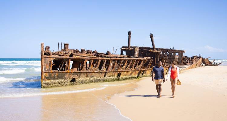 Pareja caminando junto al naufragio oxidado del Maheno en las arenas blancas de la Isla Fraser
