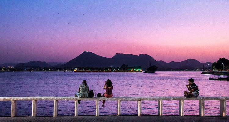 Silhouetted people sitting by a lake at sunset with mountains in the background.