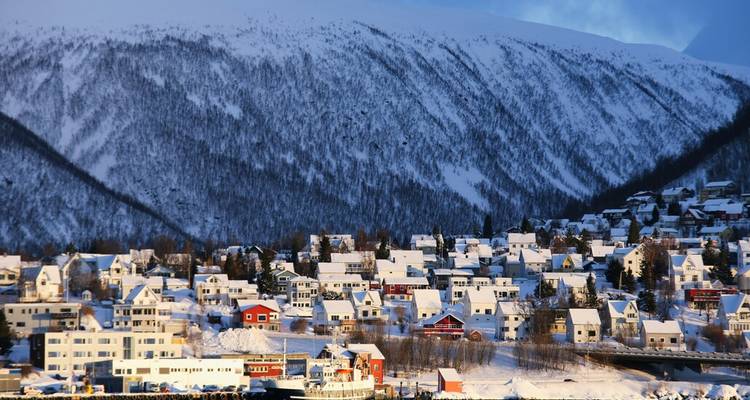 Des maisons recouvertes de neige regroupées sous une immense montagne blanche à la lumière hivernale arctique.