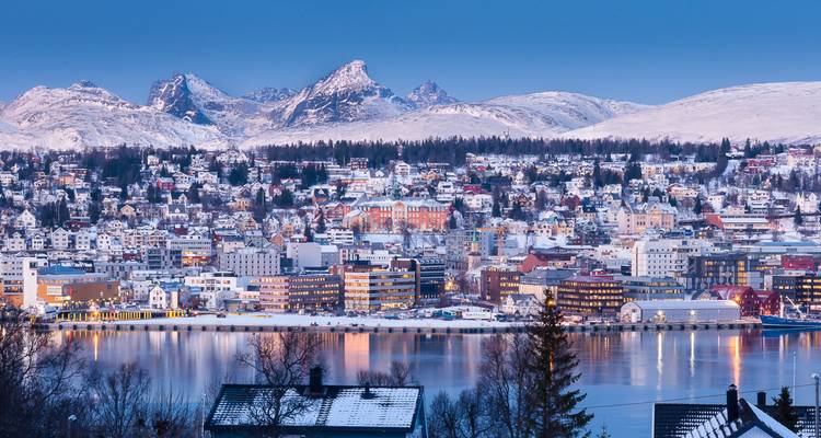 Paysage urbain hivernal panoramique avec des bâtiments couverts de neige se reflétant dans un fjord calme, encadré par des montagnes escarpées au crépuscule.
