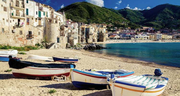 Cefalù beach with boats and historic buildings.