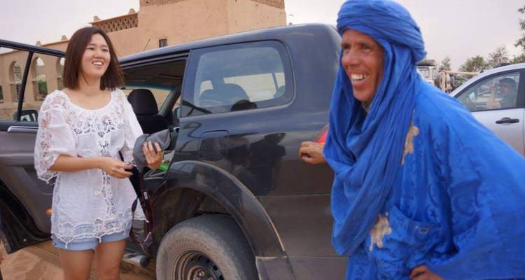 Homme et femme debout près d'une voiture avec des bâtiments du désert en arrière-plan.