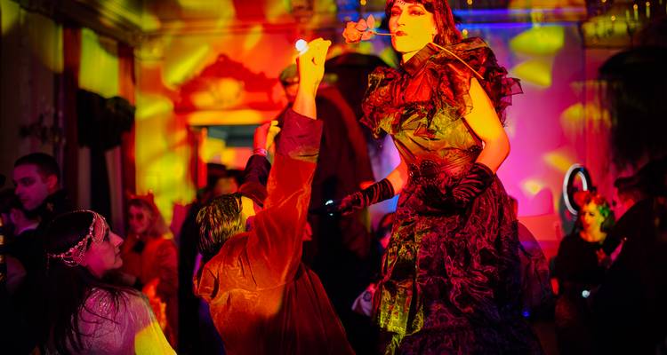 Kostümierte Frau auf Stelzen überreicht einer knienden Gästin eine Rose unter psychedelischer Beleuchtung.