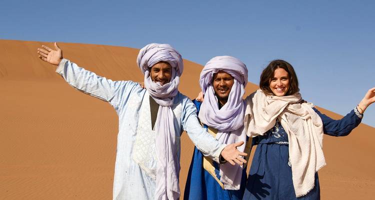 Trois personnes portant des vêtements traditionnels posent devant des dunes de sable.